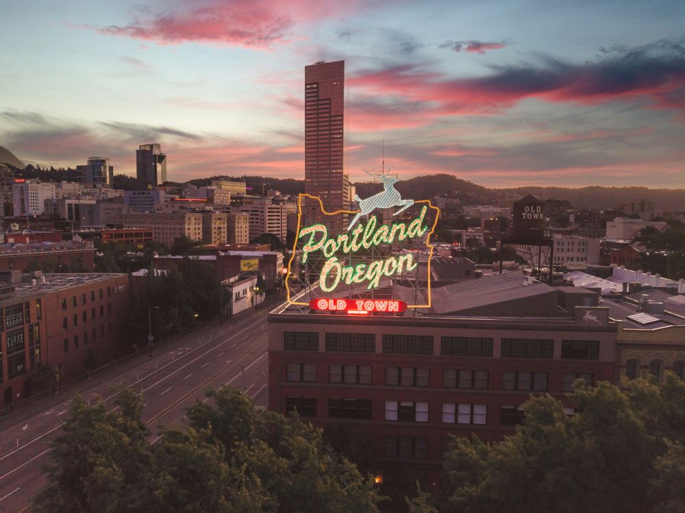 Portland, Oregon Old Town neon sign glowing at dusk with city buildings and a colorful sky in the background.