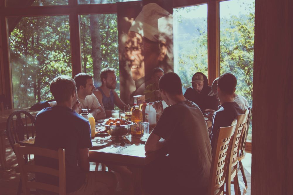 Eight people sit around a sunlit table with breakfast, drinks, and fruit, next to large windows and a mural of a smiling chef.