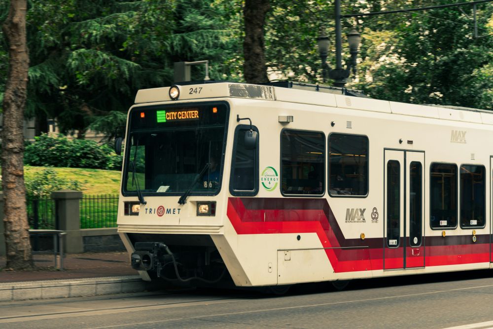 A white and red Portland MAX light rail train labeled “City Center” travels on a street lined with trees and greenery.