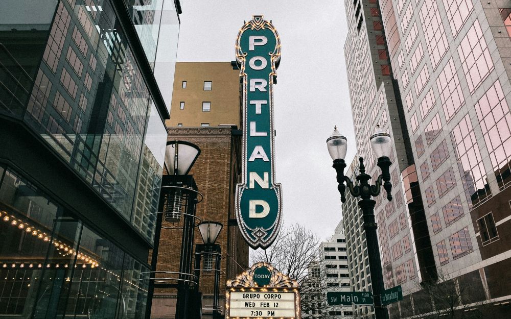 Large vertical “PORTLAND” sign on a theater, surrounded by modern buildings and streetlights on a cloudy day.